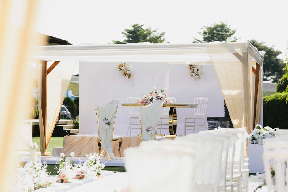 A floral arrangement scene under a cream-colored canopy featuring a round table decorated with pink and white flowers, including roses and peonies, complemented by white chairs with gold accents and f