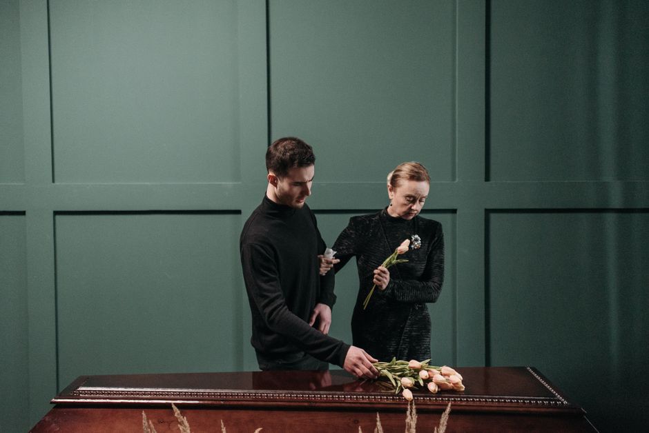 A young man and an older woman are standing at a dark wooden funeral casket with a green paneled wall behind them. The young man, dressed in a black long-sleeve shirt, is placing a light pink rose bou