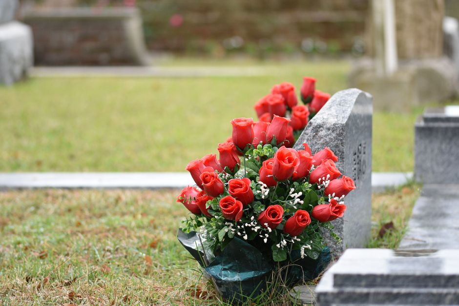 A bouquet of fresh red roses with tightly closed buds and lush green leaves, arranged with small white filler flowers, rests against a gray granite headstone in a cemetery. The bouquet is wrapped in d