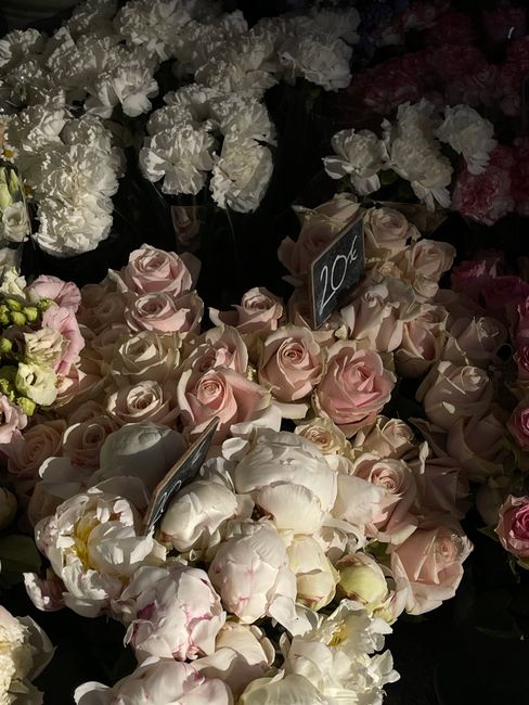 A close-up of a floral display featuring pale pink and ivory roses arranged in a bouquet, with some large white peonies and other mixed blooms nearby. The roses have delicate, softly curled petals, an