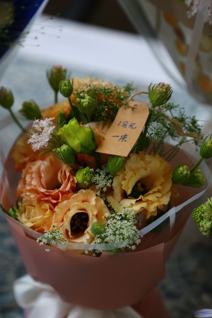 A floral arrangement featuring soft peach and cream carnations with ruffled petals, complemented by small green buds and delicate white baby's breath. The bouquet is wrapped in light pink paper and is