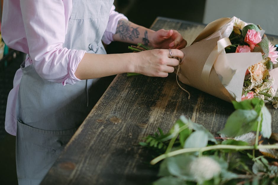 A florist in a light pink long-sleeve shirt and grey apron is arranging a bouquet of fresh flowers on a rustic wooden table. The bouquet is wrapped in light brown paper and tied with a cream ribbon, f