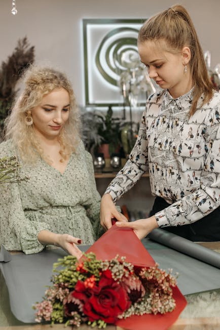 Two women are working together to prepare a floral gift in a well-lit room. The florist, with long hair tied back, is carefully wrapping a bouquet of vibrant red roses, pink baby's breath, and greener