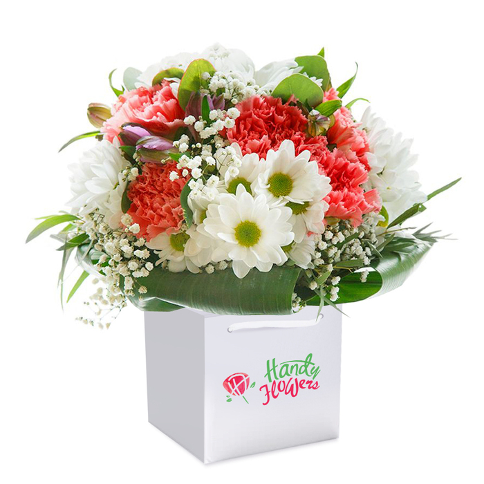 Within a pristine white square gift bag, an artful bouquet sits upright and radiant, embodying the skill of Belmont's dedicated flower artisans. The arrangement captivates with a lively blend of ruffled coral-pink and deep red carnations-texturally plush and inviting-paired with a generous scattering of elegant white daisies. These daisies, with their luminous yellow-green hearts, bring a sunny optimism to the display, recalling the friendly, open spaces of Belmont. Delicate wisps of gypsophila weave between the larger blooms, lending an airy grace, while glossy, broad-leaved greenery forms a lush cradle at the base, reminiscent of the flourishing foliage in local Belmont gardens. Sprinkled in the mix are several purple-tinged buds, possibly alstroemeria, peeking out with subtle sophistication. Every flower is meticulously placed, creating a balanced, full silhouette that holds the promise of celebration-be it for an anniversary, birthday, or any special Belmont event. The white gift bag, graceful with its soft ribbon handles, adds refined simplicity and practical elegance, ideal for seamless local delivery. Perfectly photographed against a clean, uncluttered backdrop, this composition draws focus squarely to the bouquet's vibrant colours and intricate textures. In every detail, from choice of buds to final flourish of greenery, the image reflects the pride and attentive touch of Belmont's local florists-ready to add a flourish of beauty to life's meaningful moments in the heart of the community.